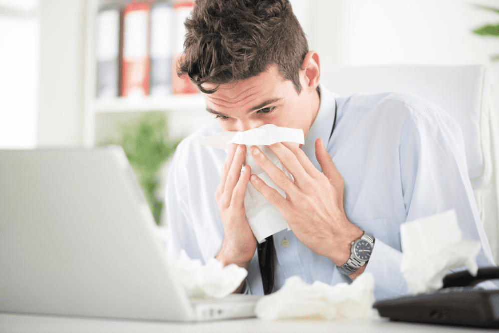 a man sitting at a desk with laptop open blowing his nose with tissues all over the desk